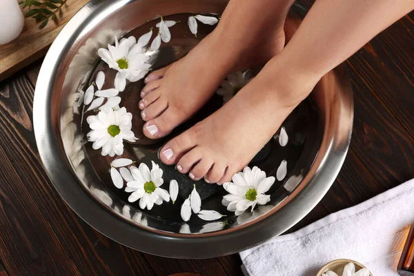 Woman soaking her feet in bowl with water, stones and flowers on wooden floor, above view. Pedicure procedure