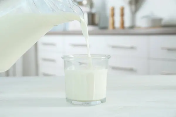 Pouring milk from jug into glass on white wooden table in kitchen