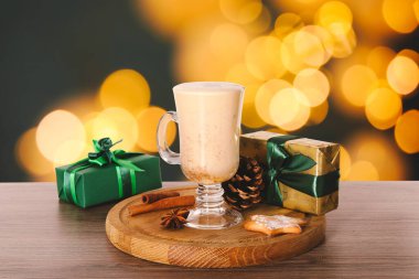 Bardaktaki nefis eggnog ve ahşap masadaki hediye kutuları bulanık Noel ışıklarına karşı