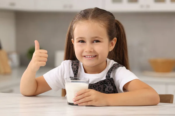 キッチンのホワイトテーブルに親指を示す新鮮なミルクのガラスとかわいい少女 — ストック写真