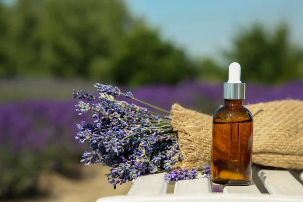 Bottle of essential oil and lavender flowers on white wooden surface outdoors, space for text