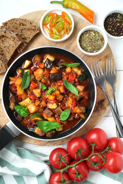 Frying pan with tasty ratatouille, ingredients and bread on white wooden table, flat lay