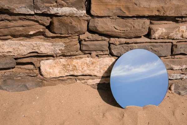 Round mirror reflecting sky on sand near stone wall outdoors, space for text