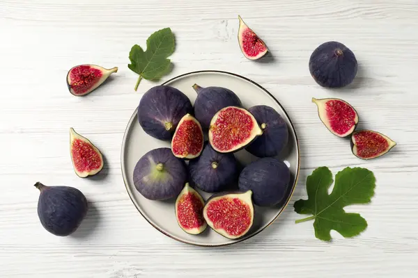 Whole and cut ripe figs with leaves on white wooden table, flat lay