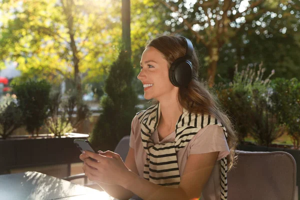 Smiling woman in headphones using smartphone in outdoor cafe. Space for text