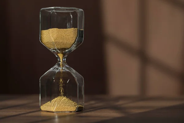 Hourglass with flowing gold sand on wooden table against brown background, space for text
