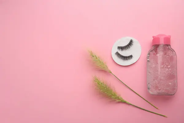 Bottle of makeup remover, cotton pad, spikelets and false eyelashes on pink background, flat lay. Space for text