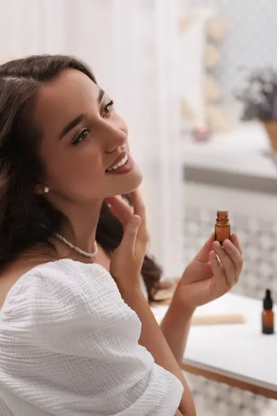 Beautiful young woman applying essential oil onto neck indoors