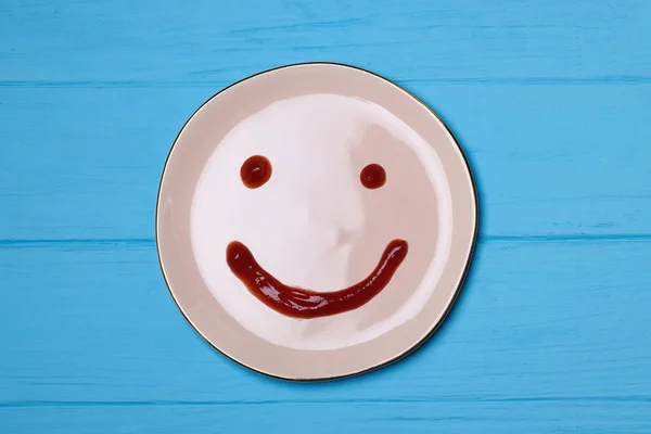 Plate with happy smiley face drawn with tomato sauce on light blue wooden table, top view