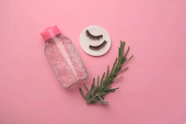 Bottle of makeup remover, cotton pad, rosemary and false eyelashes on pink background, flat lay