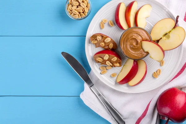 Slices of fresh apple with peanut butter, nuts and knife on light blue wooden table, flat lay. Space for text