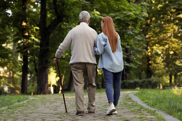 Senior man with walking cane and young woman in park, back view