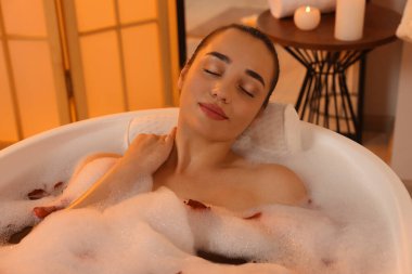 Kadın küvette köpük ve gül yapraklarıyla banyo yapıyor.