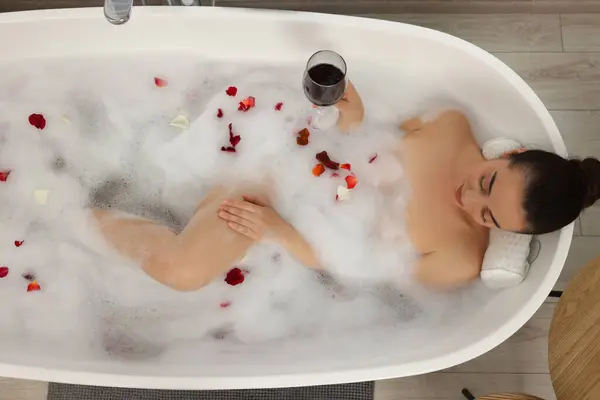 Woman with glass of wine taking bath in tub with foam and rose petals indoors, top view