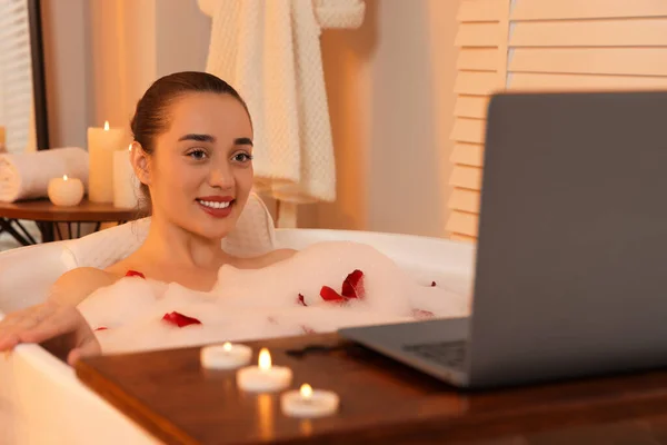 Happy woman with laptop taking bath in tub with foam and rose petals indoors