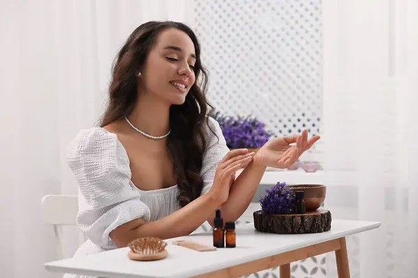 Beautiful young woman applying essential oil onto wrist at table indoors