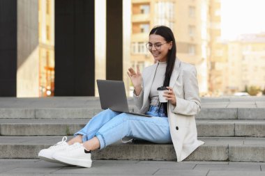 Modern bilgisayarı kullanarak merdivenlerde görüntülü konuşma yapan mutlu genç bir kadın.