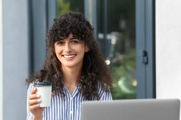 Happy young woman with cup of coffee using modern laptop outdoors