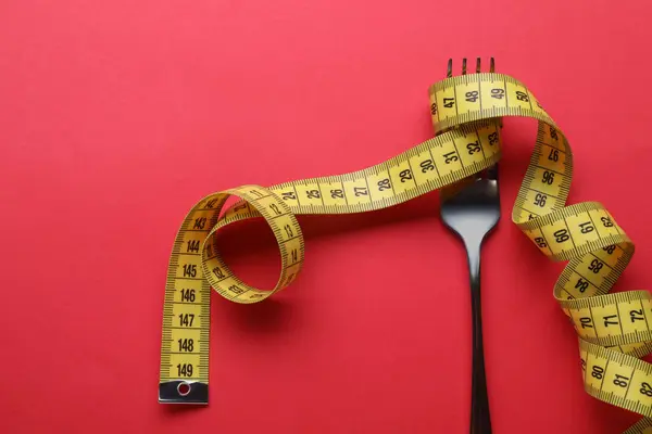 Fork with measuring tape on red background, top view. Diet concept