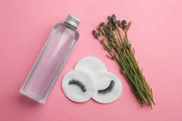 Bottle of makeup remover, cotton pads, false eyelashes and lavender on pink background, flat lay