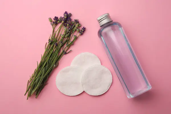 Bottle of makeup remover, cotton pads and lavender on pink background, flat lay