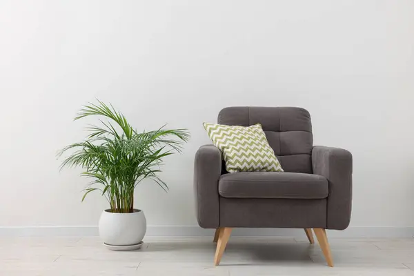 Comfortable armchair with cushion and houseplant near white wall indoors. Interior design
