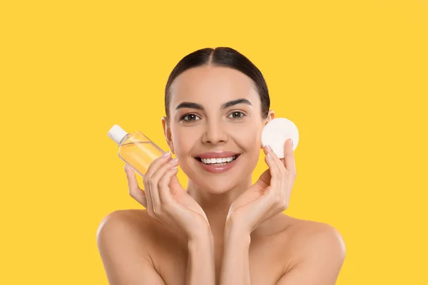 Beautiful woman holding makeup remover and cotton pad on yellow background