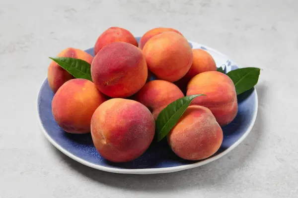 Stock image Many whole fresh ripe peaches and green leaves in plate on white table, closeup