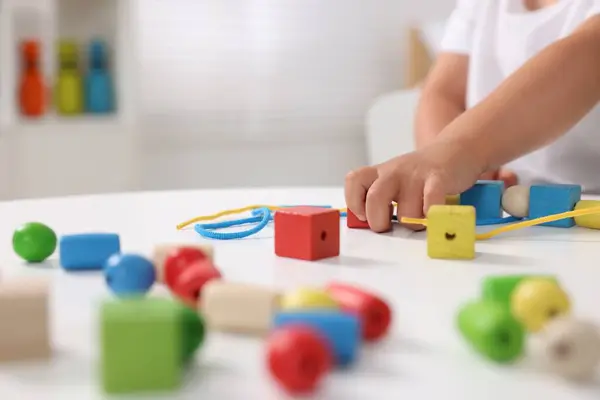 Motor skills development. Little boy playing with wooden pieces and string for threading activity at white table indoors, closeup