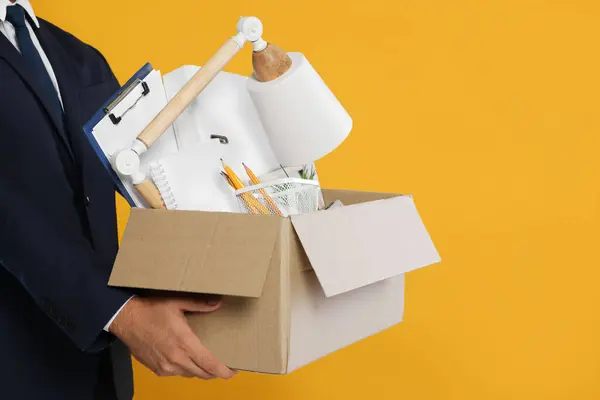 Unemployed man with box of personal office belongings on orange background, closeup. Space for text