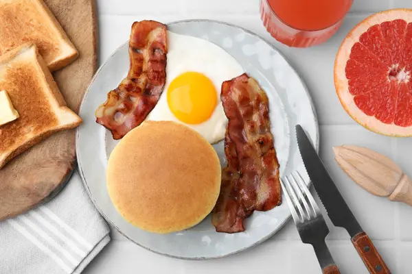 Tasty pancakes with fried egg and bacon served on white tiled table, flat lay