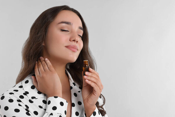 Beautiful young woman applying essential oil onto neck on light grey background, space for text