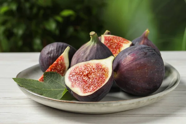 Whole and cut ripe figs with leaf on white wooden table against blurred green background, closeup