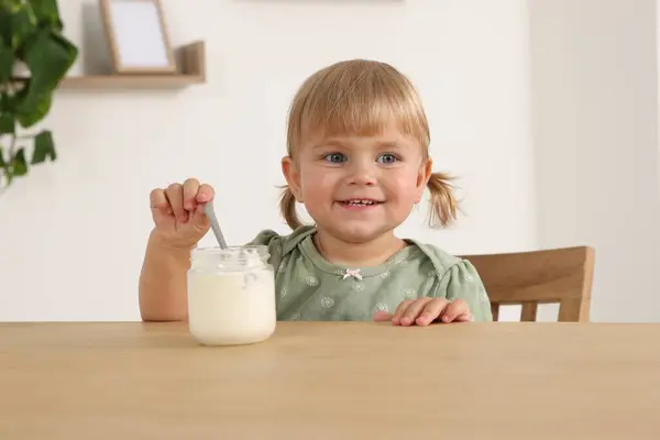 木のテーブルで瓶からスプーンで美味しいヨーグルトを食べるかわいい小さな子供 — ストック写真