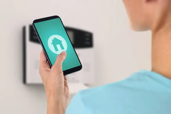 Woman operating home alarm system via mobile phone against white wall with security control panel, closeup. Application with illustration of house on device screen