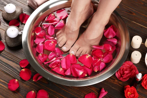 Woman soaking her feet in bowl with water and red rose petals on wooden floor, closeup. Pedicure procedure