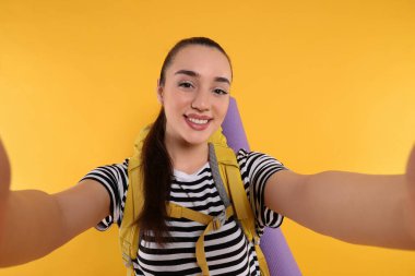 Sırt çantalı gülümseyen genç bir kadın turuncu arka planda selfie çekiyor. Etkin turizm