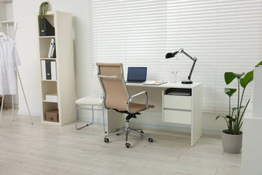 Doktorun işyeri. Klinikteki beyaz masada sandalye, dizüstü bilgisayar ve dosya.