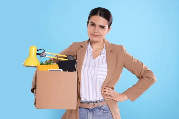 Unemployed woman with box of personal office belongings on light blue background