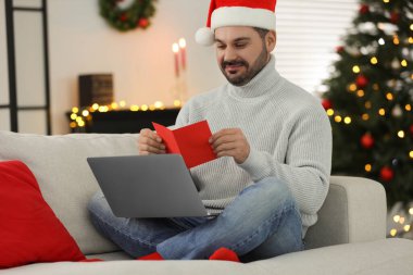 İnternette Noel 'i posta hediyeleriyle kutluyoruz. Noel Baba şapkalı bir adam evdeki bilgisayardaki video görüşmesi sırasında tebrik kartı okuyor.
