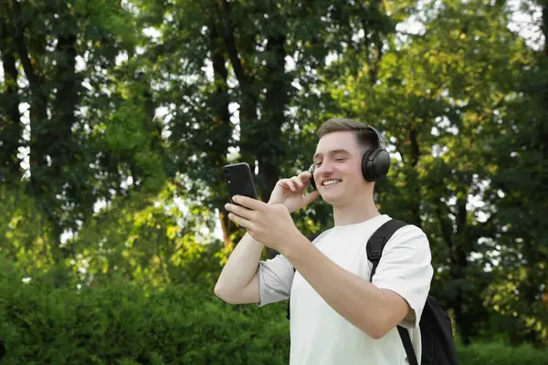 Smiling man in headphones using smartphone in park. Space for text
