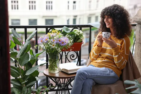 Young woman with cup of drink relaxing near green houseplants on balcony