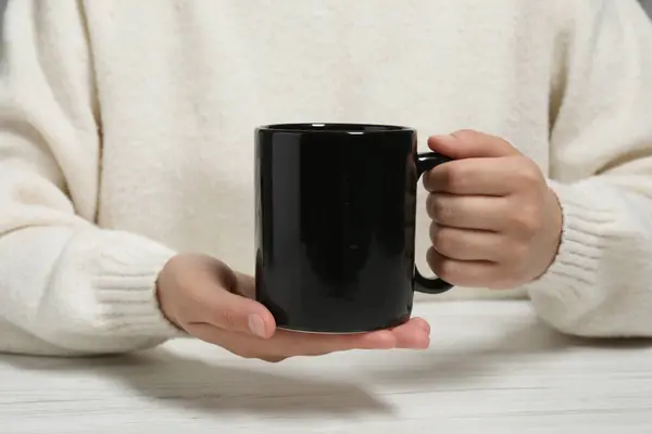 Woman holding black mug at white table, closeup. Mockup for design