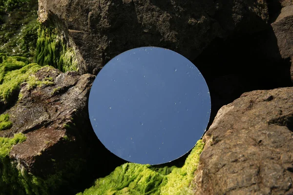 Round mirror reflecting light blue sky on stones with seaweed outdoors