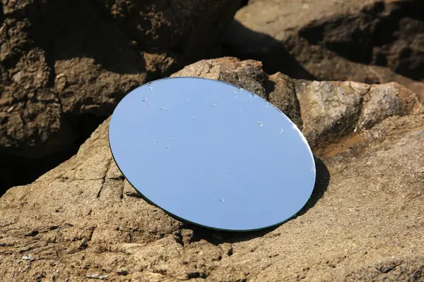 Round mirror reflecting light blue sky on stones outdoors