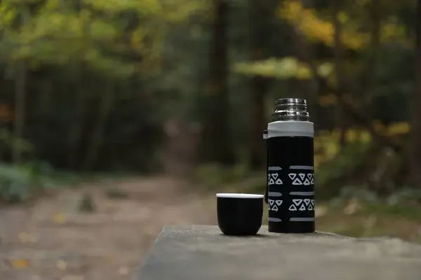 Black thermos and cup lid on wooden bench outdoors, space for text