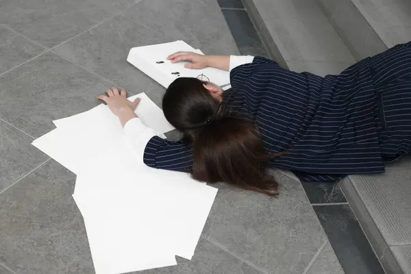 Unconscious woman with scattered folder and papers lying on floor after falling down stairs indoors