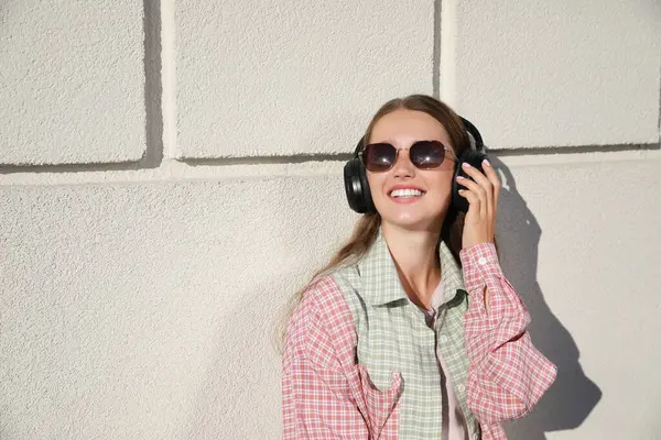 Smiling woman in headphones listening to music near white wall outdoors. Space for text