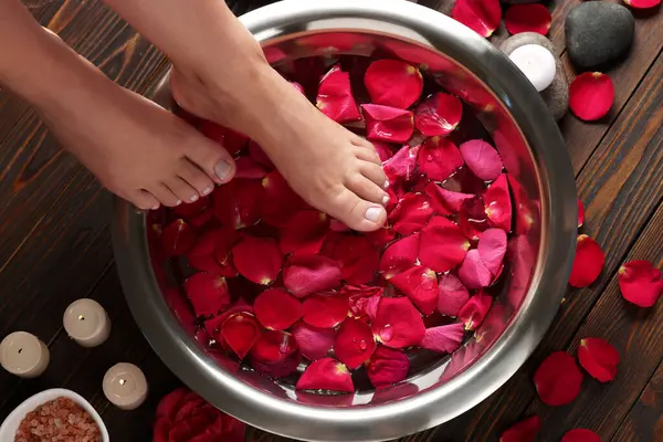 Woman soaking her feet in bowl with water and red rose petals on wooden floor, top view. Pedicure procedure