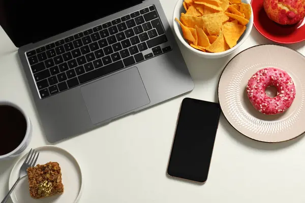 Bad eating habits at workplace. Laptop, smartphone and different snacks on white table, flat lay